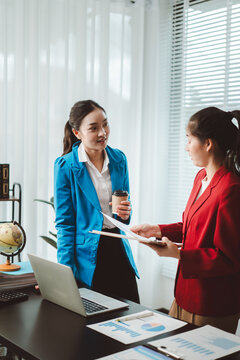 Two asian businesswomen are standing and engaging in a professional discussion, reviewing documents and charts while brainstorming ideas in a modern office setting