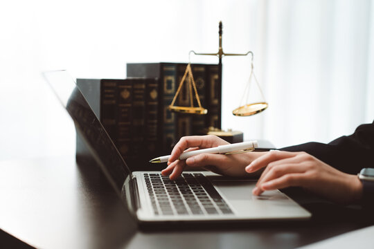 Lawyer's hands typing on a laptop and holding a pen beside legal books and golden scales of justice on a bright office desk, symbolizing legal research and advocacy