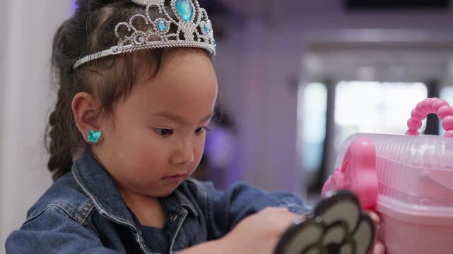 A curious toddler girl plays dress-up with a tiara and glitter makeup, exploring her imagination and developing self-expression while playing indoors.