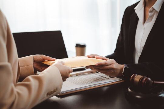 Businesswoman handing brown envelope containing document or money to a lawyer during a legal consultation, discussing contract agreement, corruption, or bribery with a gavel on the table