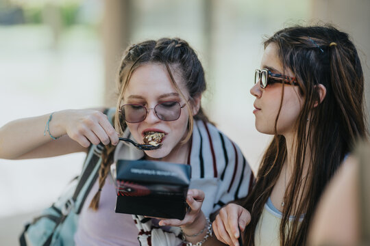 Two teenage friends sit outdoors as one eats a dessert from a takeout box with a spoon. The pair share a candid moment of casual snacking and conversation in a relaxed setting.