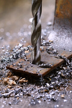 Close-up of a metal drill bit boring a hole through a rusty iron plate with metal shavings and swarf scattered around.