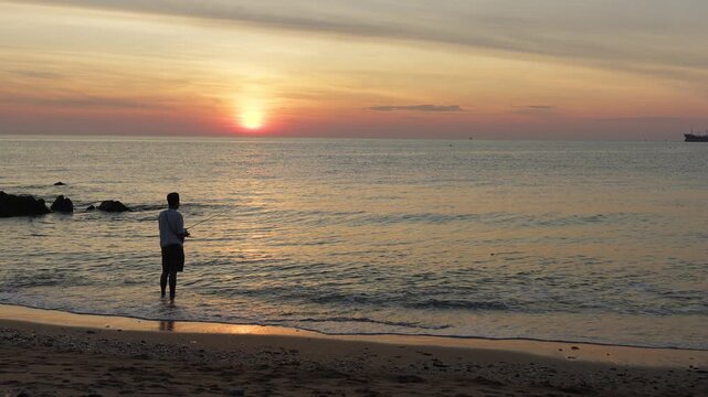 A lone fisherman stands in the shallow ocean, enjoying a tranquil morning of surf fishing as the warm sun rises beautifully over the calm sea horizon.