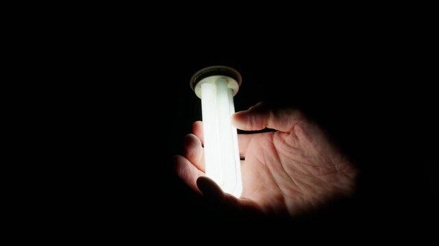 A hand gently touches a glowing energy-saving light bulb against a pitch-black background. This illustrates the concept of cool-to-the-touch energy efficiency and modern power saving.