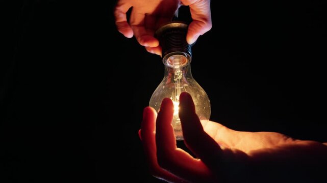 Two hands gently screw an incandescent light bulb into a socket. The delicate internal filament is visible against a solid black background. The surrounding area remains completely dark.