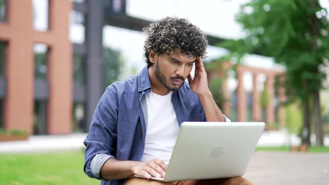 Worried male student read bad news on laptop while sitting on a bench in campus space near the university building. Frustrated stressed handsome man upset looking at message on computer. Close up
