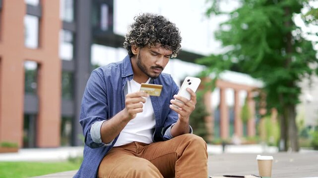 Frustrated stressed young man detected fraud while entering credit card data on a mobile phone sitting on bench on the street. Worried male became a victim of fraud, money was stolen from his account