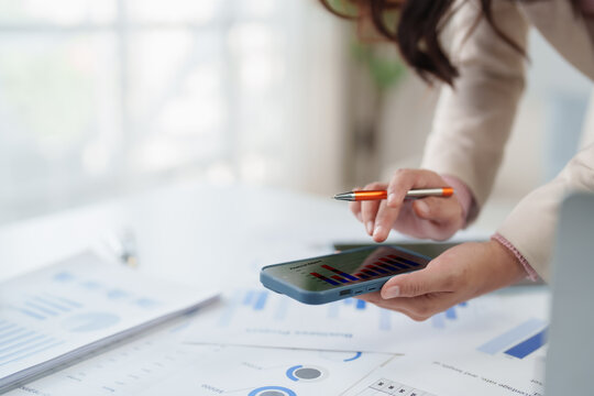 Person holding a smartphone, analyzing financial data with bar graphs on the screen, comparing business reports and charts on a desk during market research
