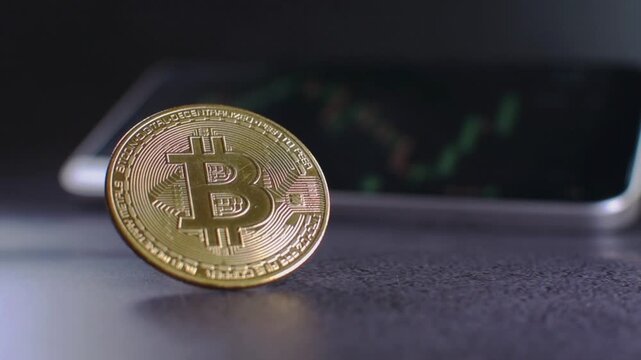 Vertical close up video of a golden bitcoin cryptocurrency coin standing on a granite surface while a blurred smartphone in the background shows a financial trading chart, symbolizing blockchain techn