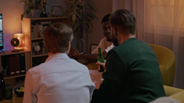 Pull out shot of biracial best friends gathering in living room, drinking beer and discussing football match, sharing emotions