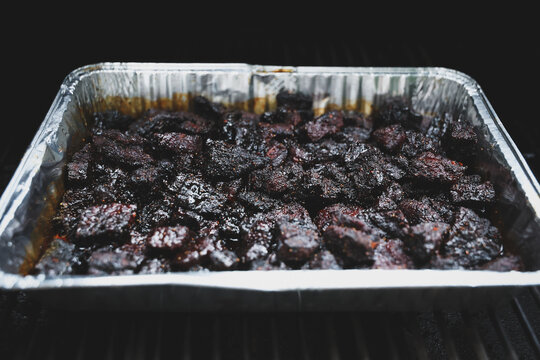 Pork Belly Burnt Ends cooked until black on a pellet grill smoker. The delicious morsels of fatty pig meat are resting in a tinfoil pan ready to eat for dinner. 