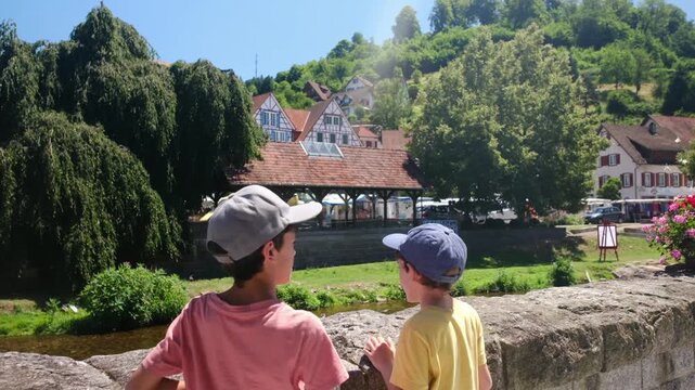 Two young brothers enjoying a scenic view of a historic german village from a stone bridge, admiring the traditional half-timbered houses and lush green landscape