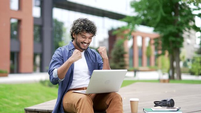 Happy excited male student received great news on laptop sitting on a bench in campus space near university building. Smiling shocked surprised handsome man reads positive good message on the computer