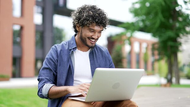 Satisfied happy male student rejoices at positive results in work on laptop while sitting on a bench in campus space near university building. Smiling joyful man received great news on the computer