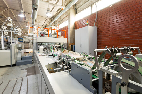 View of a folder gluer machinery system in operation at a printing plant for the creation of paper bags including various rollers and belts for a fast and automated assembly process in the factory.