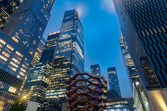 Looking up at the tall and modern skyscrapers of the Hudson Yards real estate development in New York during the evening with illuminated windows and the Vessel in the foreground.