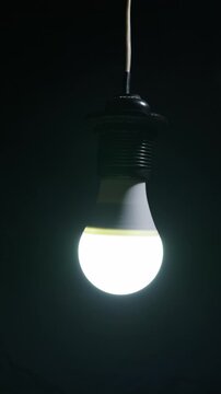 A glowing LED light bulb hangs from a wire in total darkness. The bright white light creates a stark contrast against the pitch-black background.