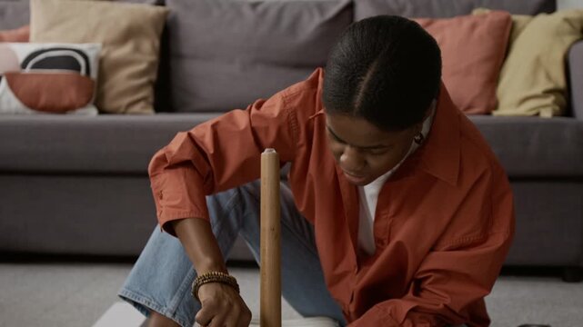 Tilt up view of Black gen Z woman screwing brown table leg to its white top while assembling furniture with paper manual