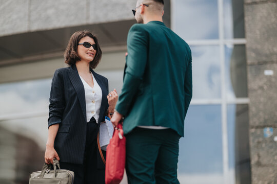 Two business people stand and converse outside a modern office building, dressed in professional attire. A brief, casual conversation between colleagues holding bags and briefcases.
