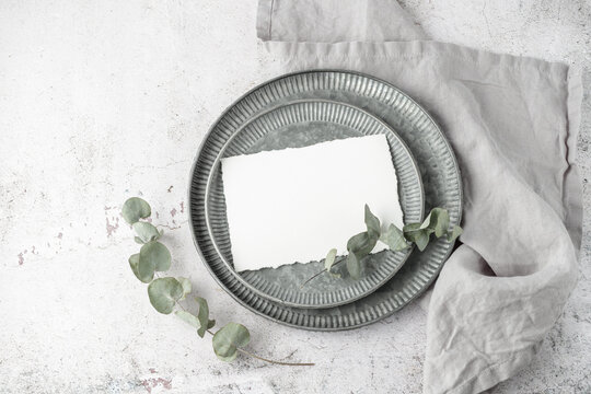 Blank white paper postcard on retro metallic round tray, eucalyptus twig and grey linen napkin on grunge stone background. Invitation floral card in vintage style. Mockup birthday greeting card.