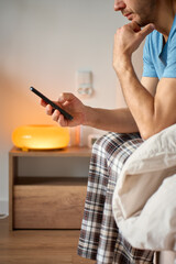 Naklejka premium Man sits on a bed in pajamas and looks at a smartphone after waking up in a bedroom interior. Concept of digital addiction, morning routine and online dependence