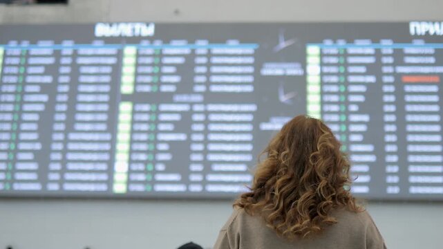 Woman stands before airport departure board. Hair flows down her back. Screen shows flight status in Cyrillic. She watches schedule intently. No animals or plants visible
