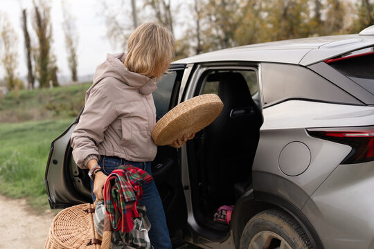 Woman car picnic preparing for road trip loading wicker baskets into silver SUV on rural path