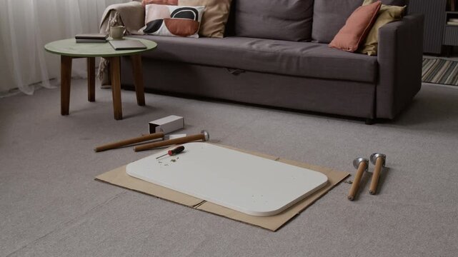 No people view of assembly tools including screwdriver, white table top and wooden legs on grey carpet, with couch and green coffee table in background