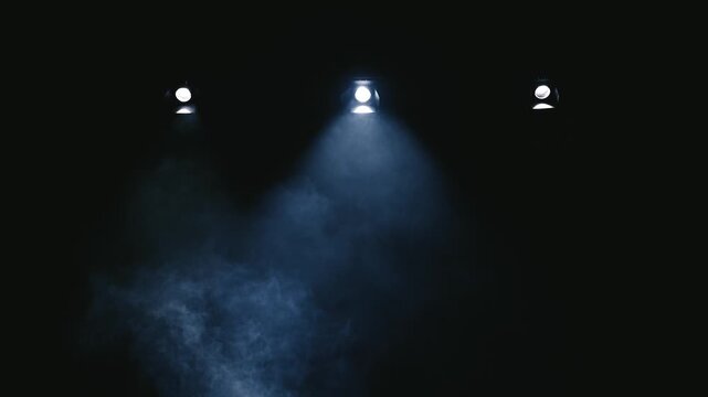 Symmetrical horizontal shot of three cold light projectors on a dark ceiling. Heavy theatrical smoke drifts and dances through the sharp light beams on an empty theater stage