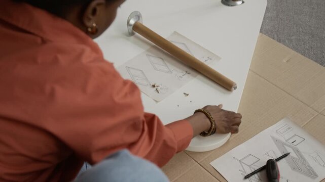 Over the shoulder view of Black girl in orange shirt turning white table top with intent to attach fourth leg to it while assembling furniture piece on cardboard sheet