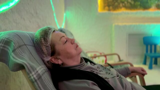 Elderly woman relaxing in salt cave halotherapy chamber, wearing cap and reclining on wooden chair with footstool, soft neon wave