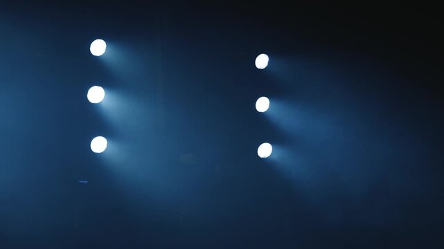 Six intense cold blue spotlights arranged in two parallel vertical rows on a dark stage. Heavy theatrical smoke and haze drift through the bright light beams