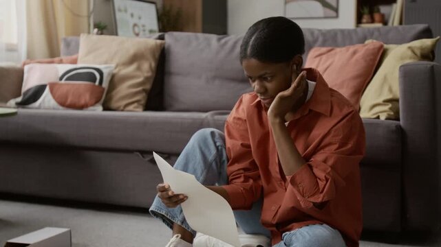 Tilt up shot of African American gen Z girl pulling up sleeves and reading instruction manual before assembling coffee table on floor in living room