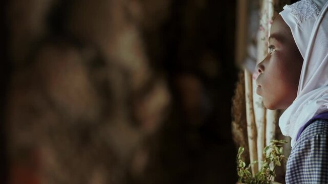 A young girl in a white headscarf looks outside through an old barred window, reflecting childhood, education, rural home life, quiet hope, innocence and thoughtful curiosity indoors.