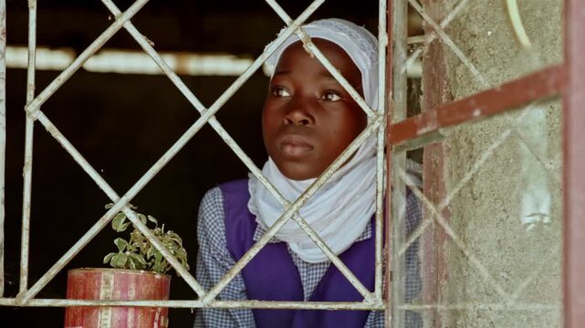 A young girl in a white headscarf looks through an old barred window beside a small potted plant, evoking childhood, hope, education, rural life, resilience and quiet curiosity indoors.