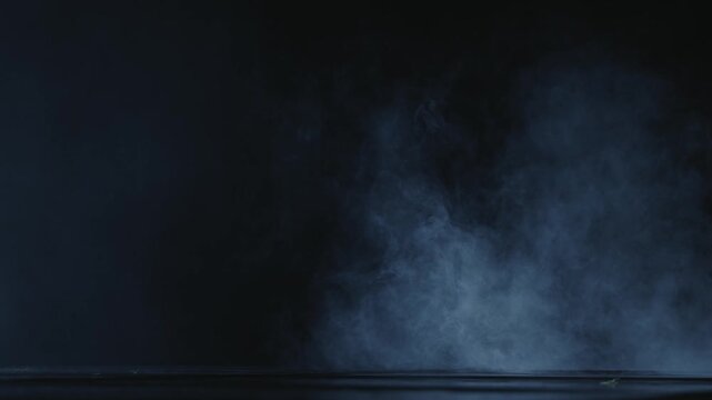High-contrast static shot from a low angle showing atmospheric theatrical fog and smoke moving slowly across a dark stage floor. Moody cold blue tint with ample negative space
