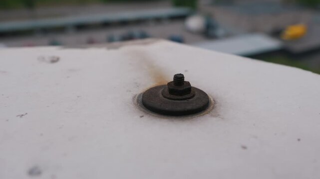 A close-up view shows a heavily rusted metal bolt, nut, and washer on a white weathered surface. A brown rust stain trails across the ledge against a soft bokeh industrial background.
