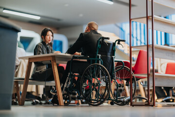 Naklejka na ściany i meble A business meeting with a man in a wheelchair speaking to a colleague at a modern office table. Colleagues sit and communicate in an accessible, collaborative workplace setting.
