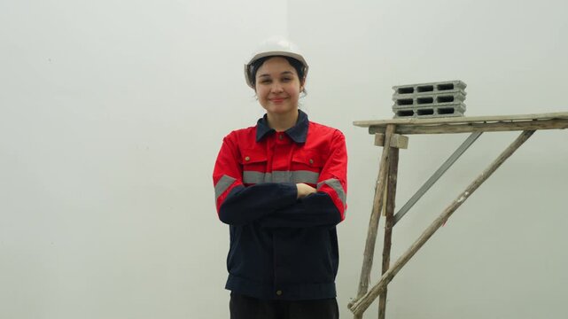 Female worker in safety helmet at construction site with crossed arms, near scaffold and concrete block, plain wall background, safety uniform, hard hat, calm expression