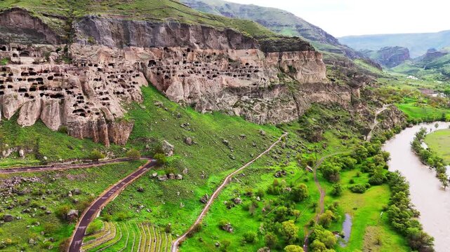 Wide aerial view of Vardzia cave monastery complex and Mtkvari River canyon, Georgia. Panoramic drone shot of the ancient rock-hewn city carved into cliffs among green spring mountains