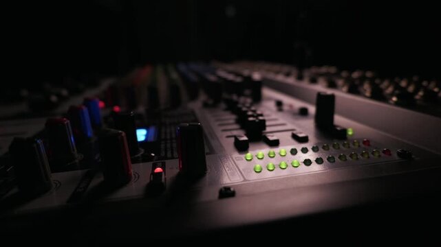 Audio sound mixing console sits in a dark recording studio with glowing green LED level meters highlighting the complex knobs and fader sliders. Concept of professional music production.