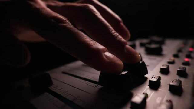 A person hand moves a fader control upward on an audio mixing desk. The dark studio setting features blurred lights in the background. Close-up details of the mixer include numerical markings.