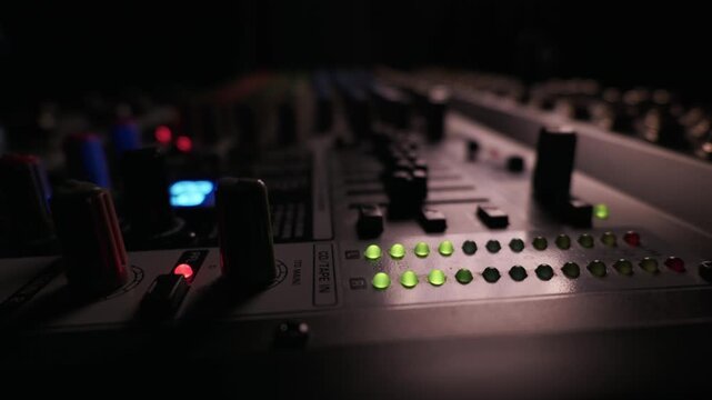 Audio mixing board features green and yellow LED volume indicators in a dark environment. Several control knobs and faders are visible with shallow focus. Red light illuminates a PFL button.