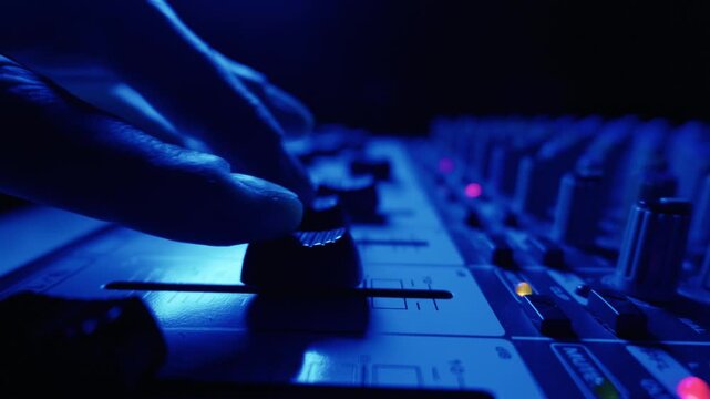 An audio engineer hand adjusts the volume faders on a professional sound mixing console. The close-up shot features illuminated buttons and knobs in a dark studio with moody cyan lighting.