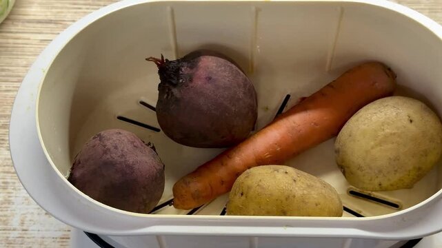 Potatoes carrots beets in steamer tray, household electric steamer without lid, closeup of unpeeled root vegetables on steaming
