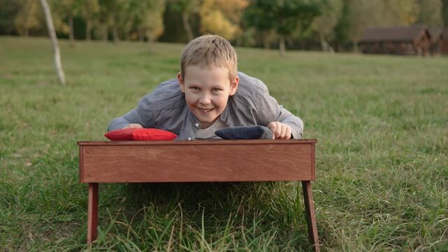 Caucasian boy lies on cornhole board and happily fools around with bean bags on camera. Portrait of happy child with colorful bags in hands sliding on board.