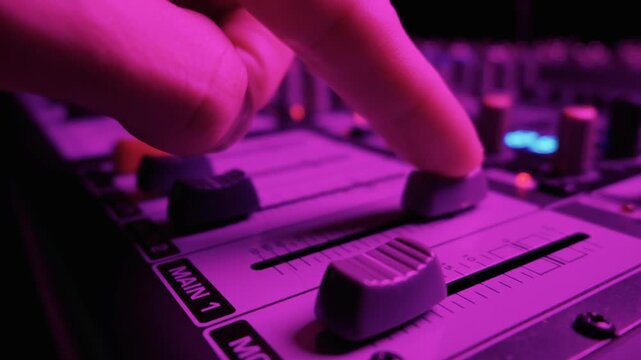 Close up of an audio mixing console featuring channel faders in a studio. The professional soundboard is illuminated by blue and red LED lights. The sliders adjust volume levels.