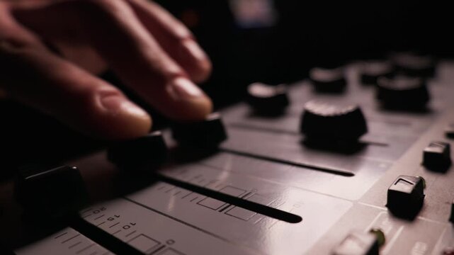 A finger pulls down a fader knob on an audio mixing console to decrease the volume. Close-up details highlight the ribbed plastic sliders. Dark studio lighting surrounds the soundboard.