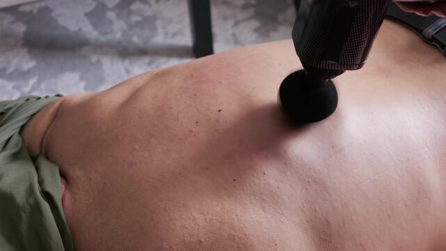 A black percussion massager works on the bare skin of a person torso to relieve tension. Concept of deep tissue therapy, sports muscle recovery, and at-home physical wellness.