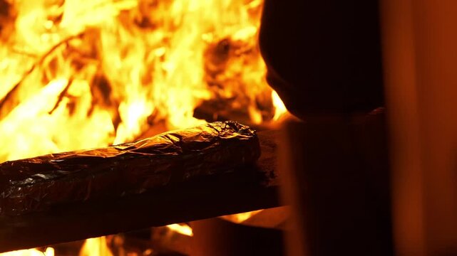 A close-up shot shows a pack of traditional prefou garlic bread wrapped in aluminum foil being carefully flipped over and placed close to the glowing embers of a campfire. The warm light of the flames